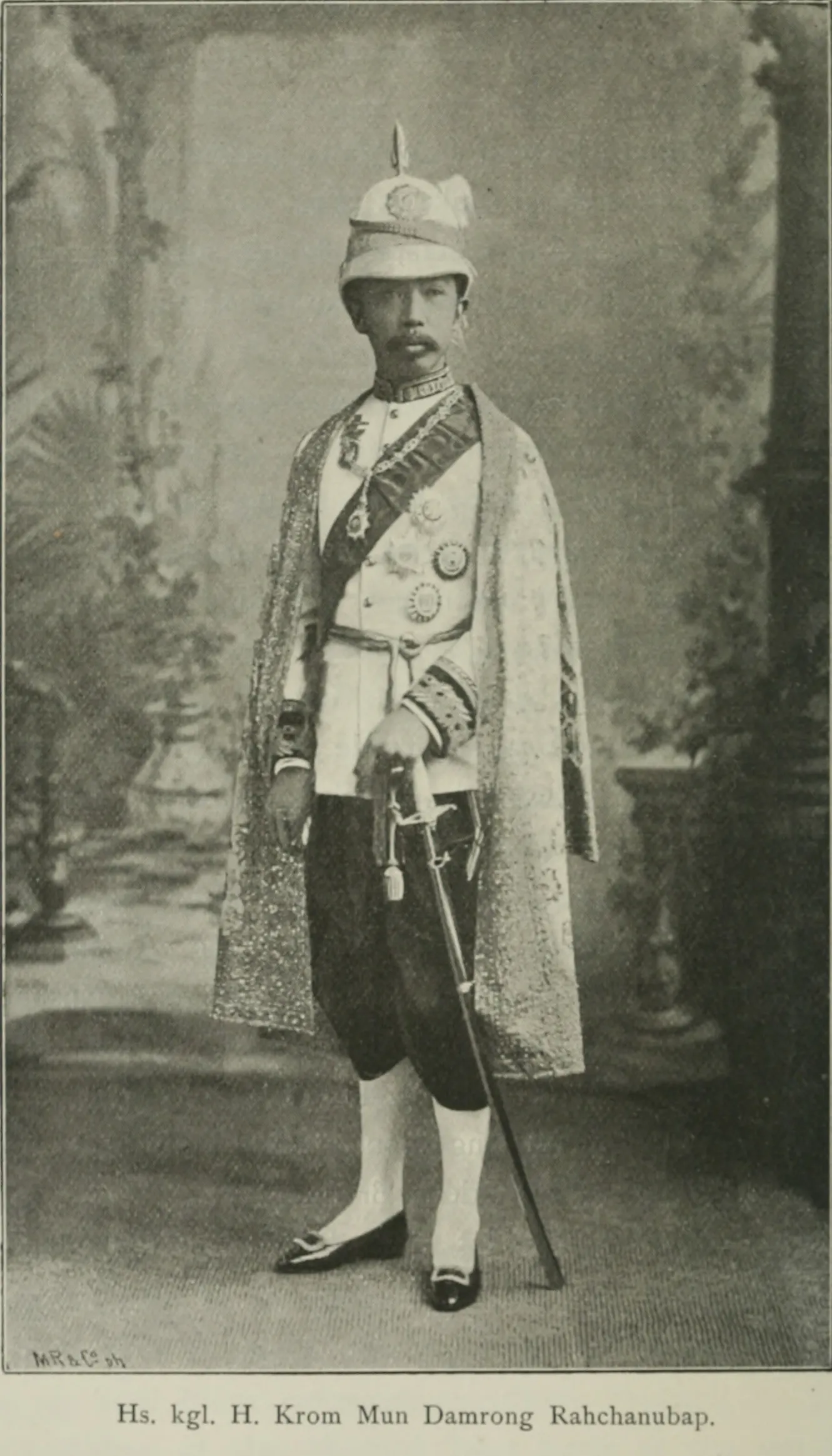 Fotografía del príncipe Damrong joven, de pie en traje de corte, con capa, casco con penacho y espada.