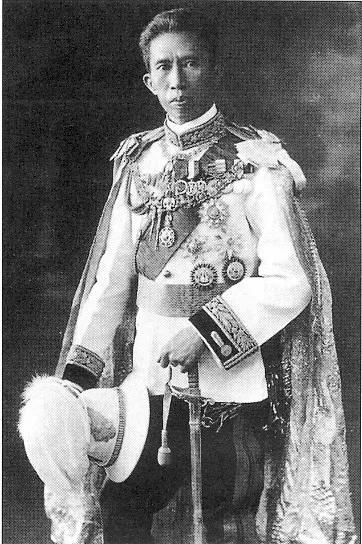 Photographie du prince Naris en uniforme blanc décoré de médailles, tenant un casque à plumet.